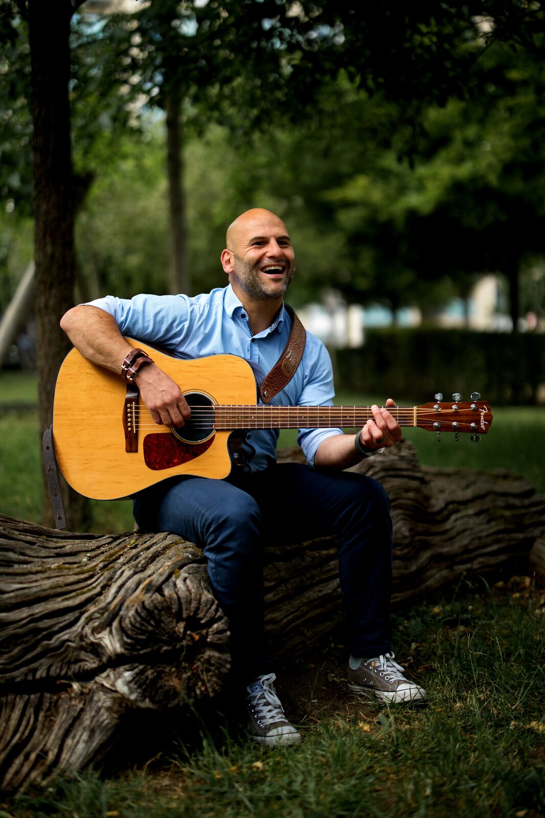 Barry de Jager seated on a fallen tree in a leafy park, laughing warmly, acoustic guitar in hand — escape became expression