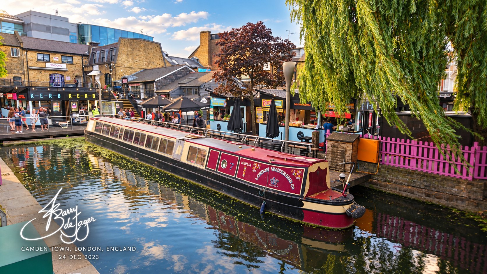 A red London Waterbus Co narrowboat moored on the canal beside market stalls and a weeping willow in Camden Town