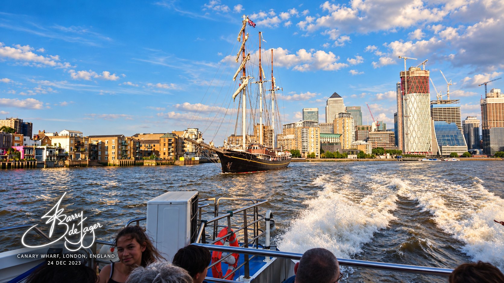 A tall ship sailing up the Thames towards the Canary Wharf towers, seen from the wake of a river boat