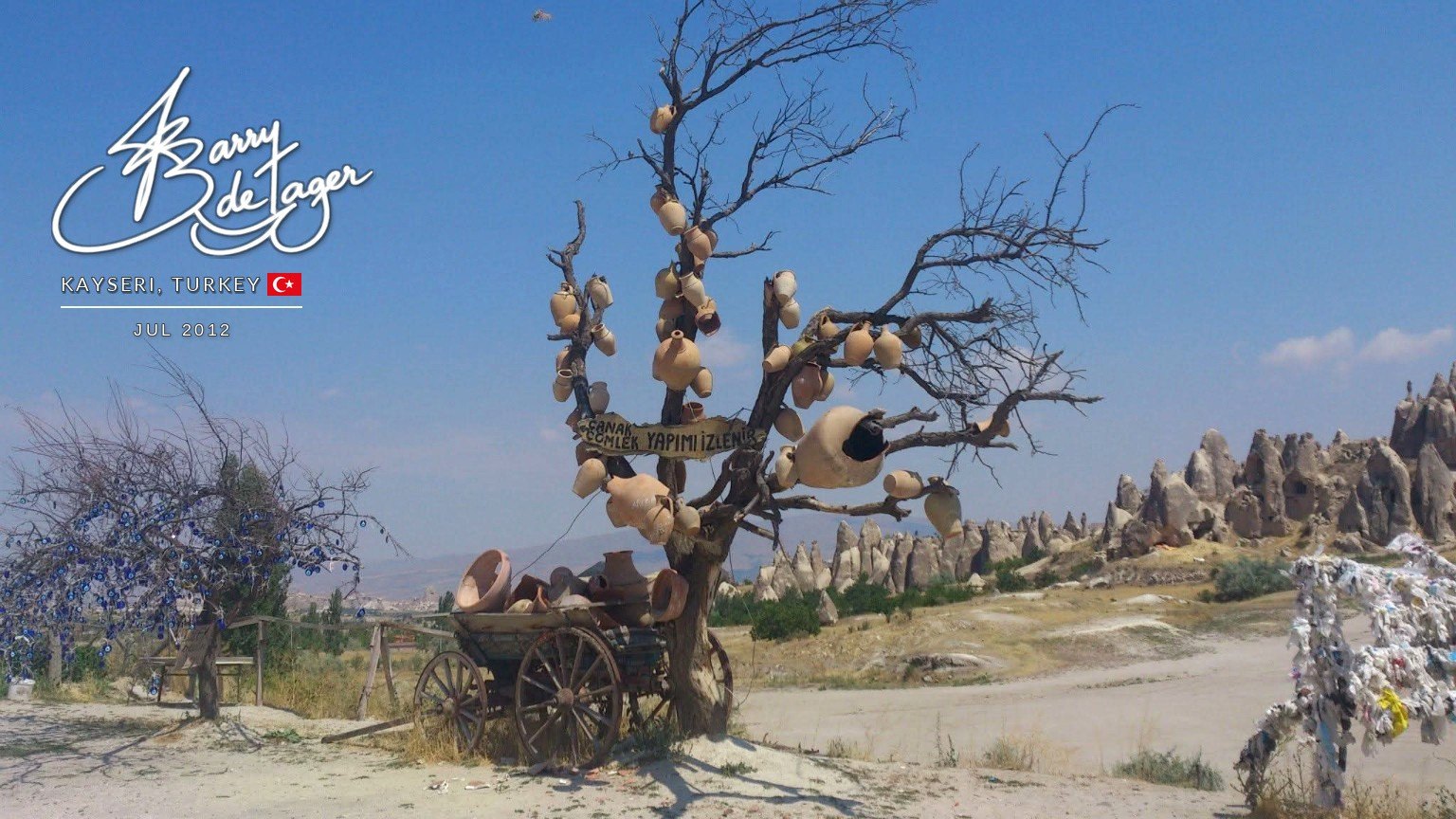 Bare tree hung with clay pots beside an old wooden cart, with fairy-chimney rock formations behind in Cappadocia
