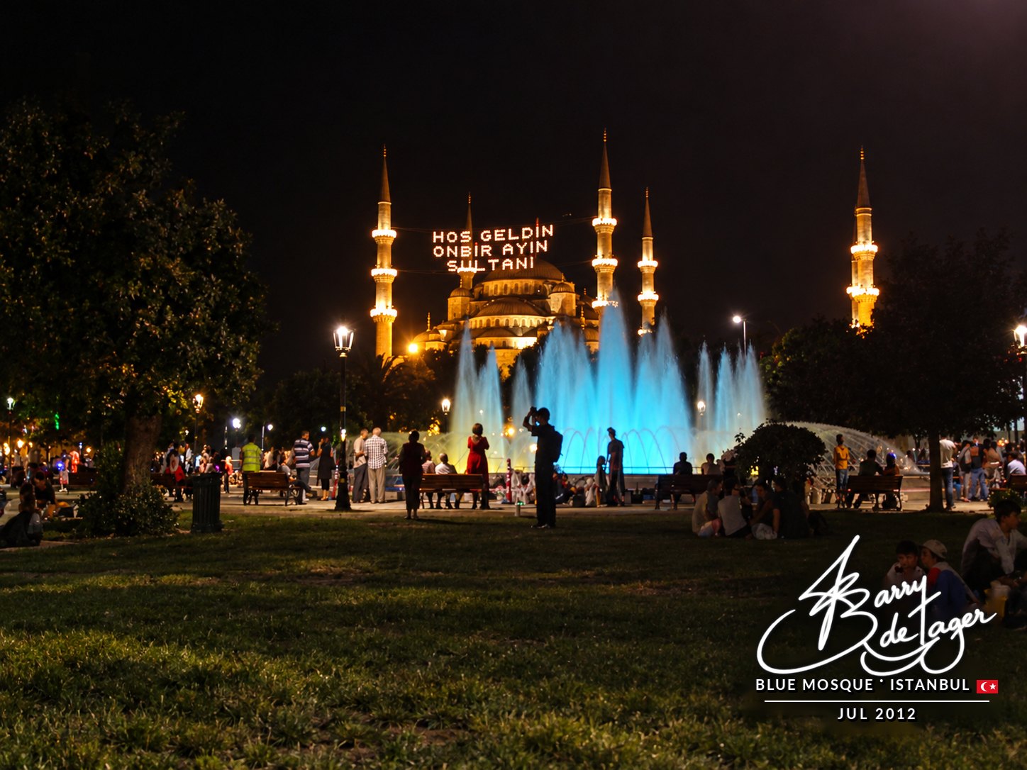 The Blue Mosque lit up at night behind illuminated blue fountains, with people relaxing on the lawn