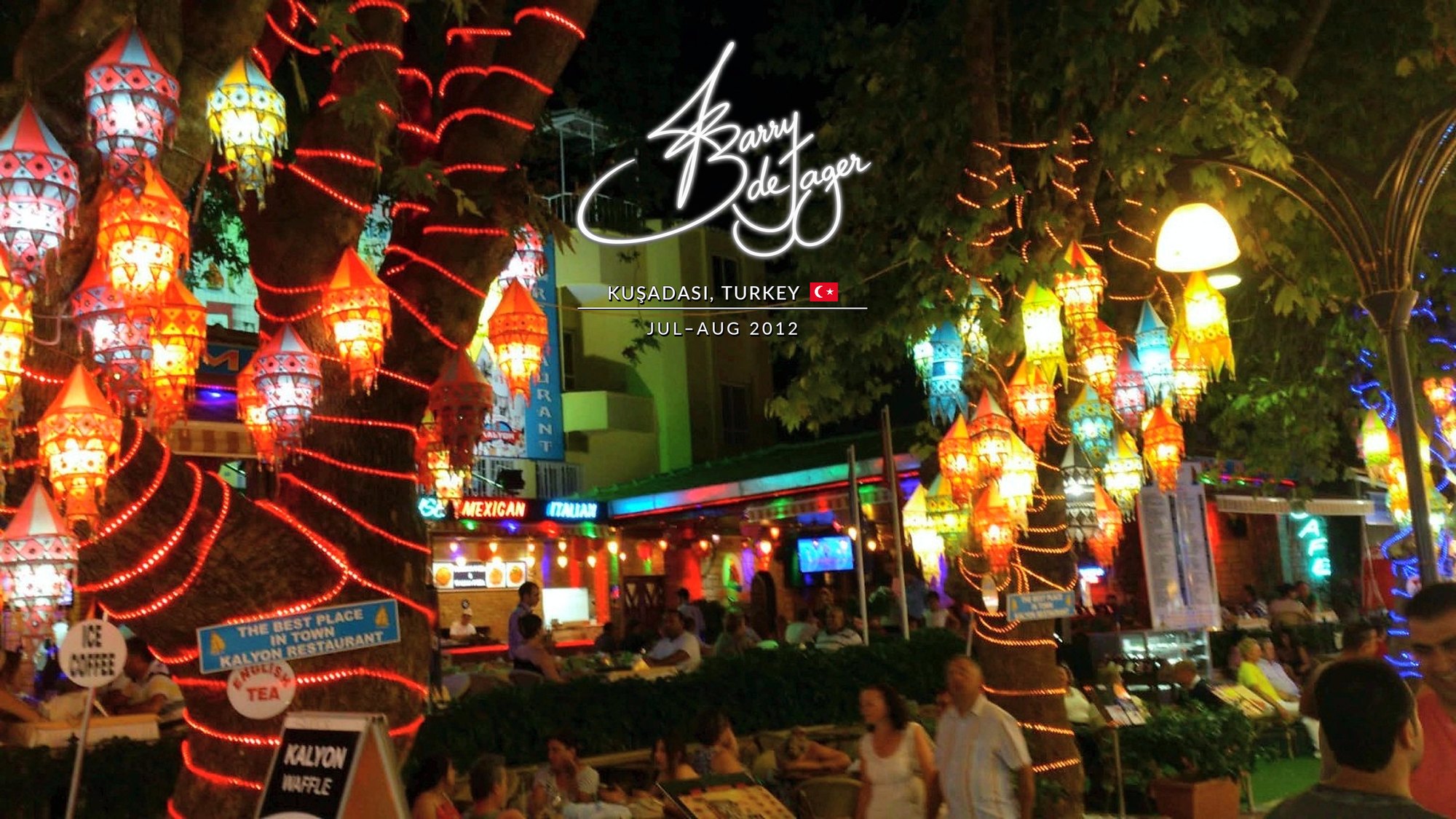 Trees wrapped in red lights and hung with glowing lanterns above busy restaurant terraces at night in Kuşadası