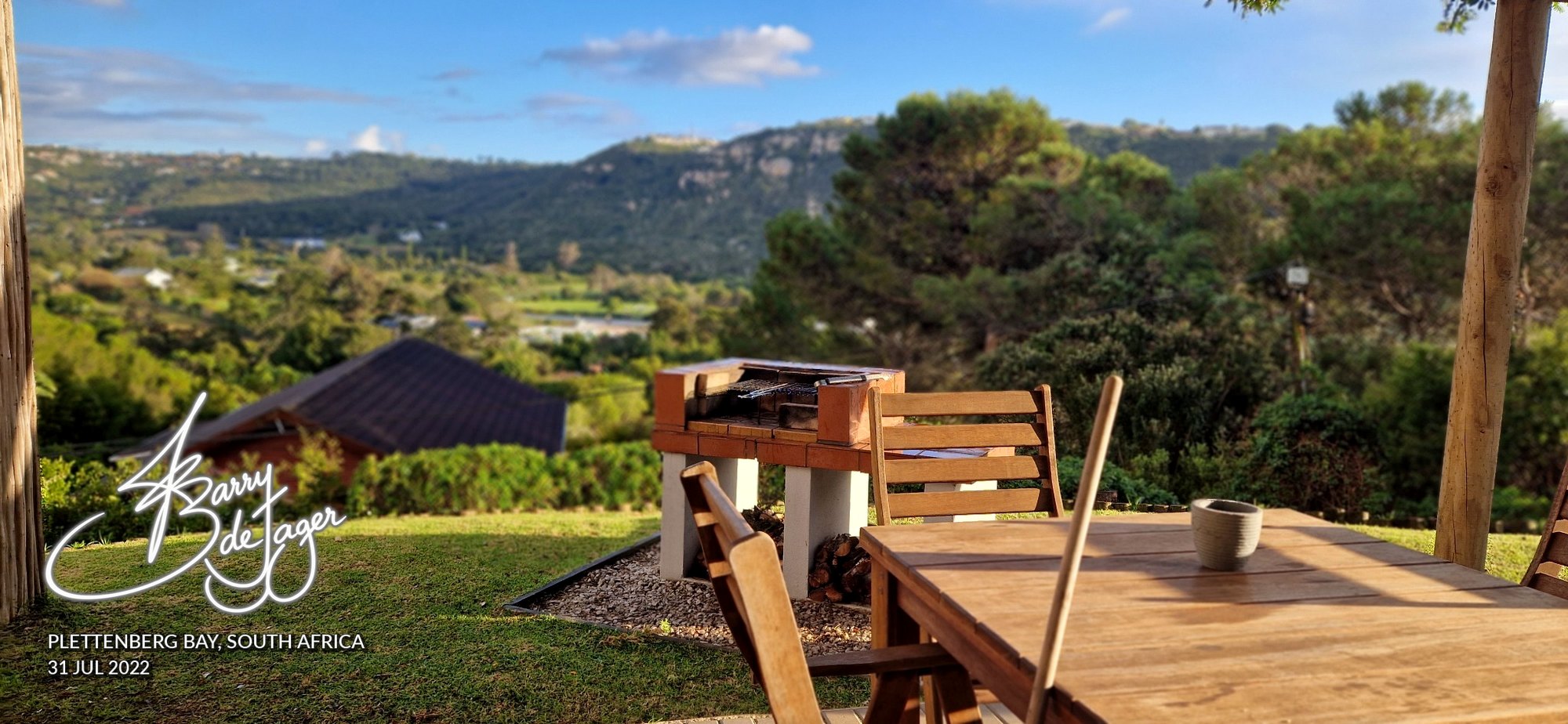Wooden table and braai on a sunny garden terrace overlooking green hills at Plettenberg Bay